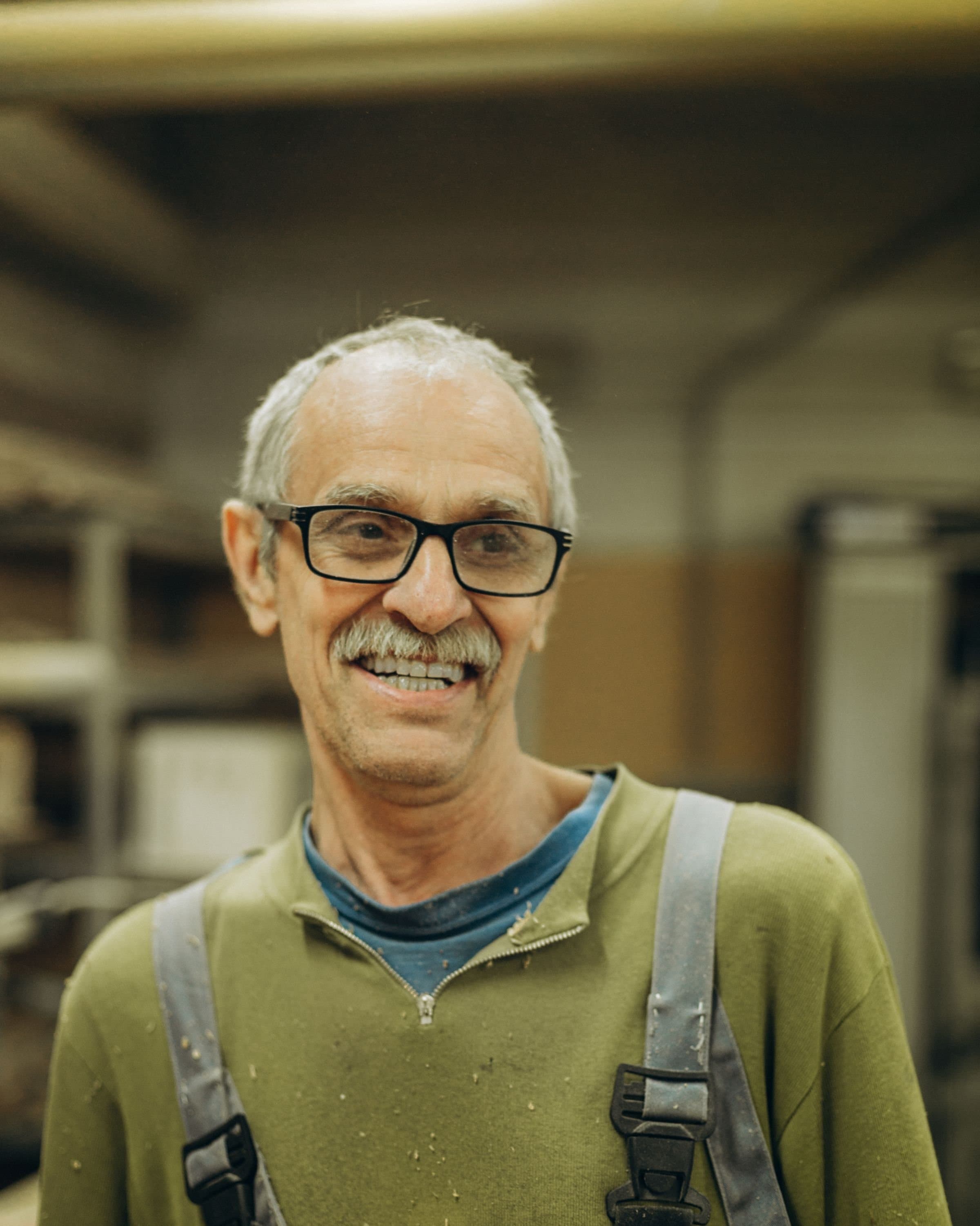 A smiling craftsman in work overalls, sawdust on his sweater, in a woodworking shop.