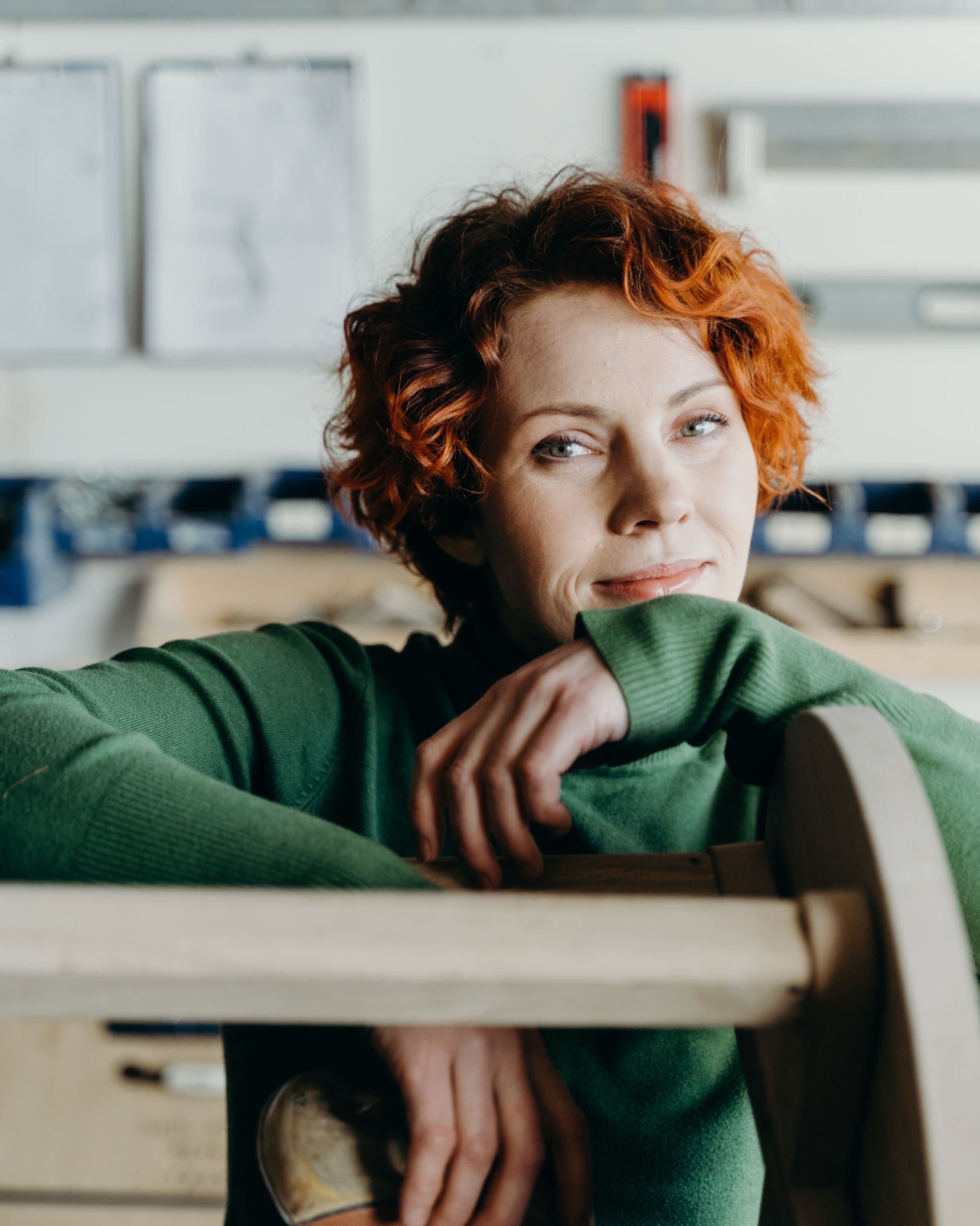 A woodworker in a green sweater leans on a chair frame in her workshop.