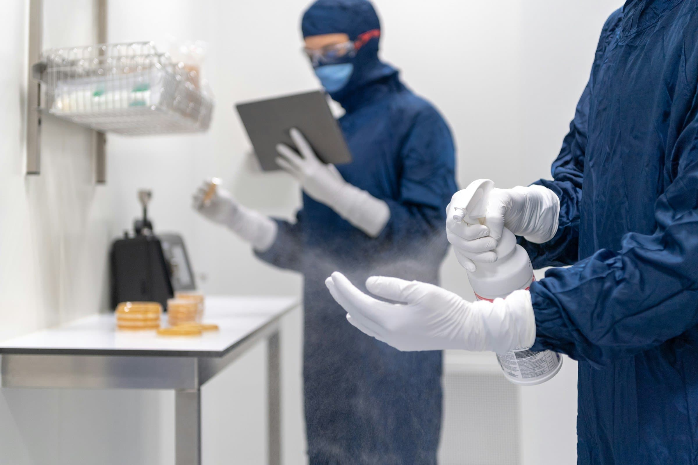 Two technicians in navy cleanroom gowns disinfect their gloves while one logs the work on a tablet.